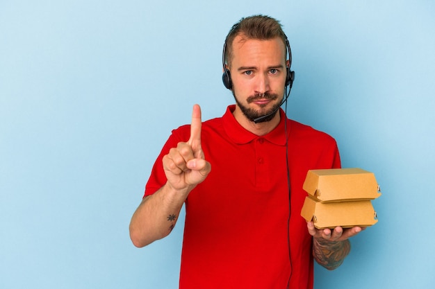 Jonge blanke bezorger met tatoeages met hamburgers geïsoleerd op een blauwe achtergrond met nummer één met vinger.