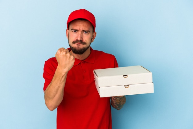 Jonge blanke bezorger met tatoeages met pizza's geïsoleerd op blauwe achtergrond met vuist naar camera, agressieve gezichtsuitdrukking.