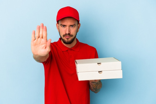 Jonge blanke bezorger met tatoeages met pizza's geïsoleerd op een blauwe achtergrond, staande met uitgestrekte hand met stopbord, waardoor je wordt voorkomen.