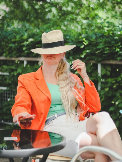 Jonge blanke blonde vrouw een feloranje jas en een strohoed zit in een openluchtcafé