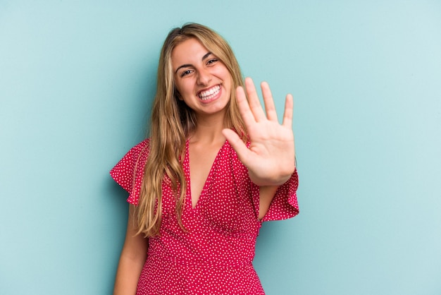 Jonge blanke blonde vrouw geïsoleerd op blauwe achtergrond glimlachend vrolijk weergegeven: nummer vijf met vingers.