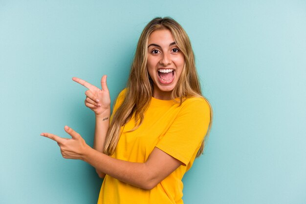 Jonge blanke blonde vrouw geïsoleerd op blauwe achtergrond opgewonden wijzend met wijsvingers weg.