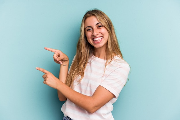 Jonge blanke blonde vrouw geïsoleerd op blauwe achtergrond opgewonden wijzend met wijsvingers weg.