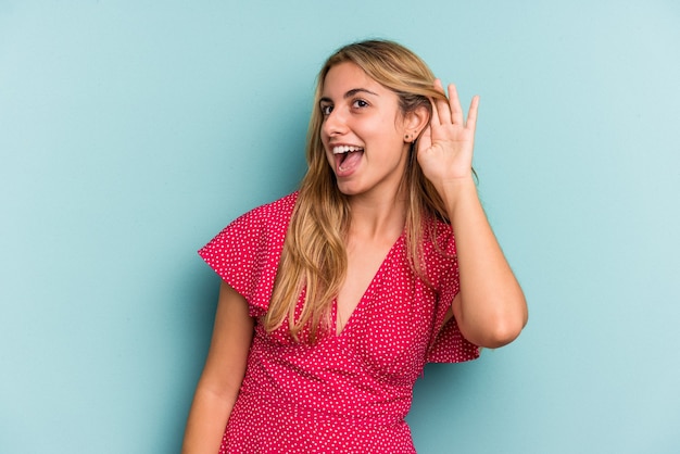 Jonge blanke blonde vrouw geïsoleerd op blauwe achtergrond probeert te luisteren naar een roddel.