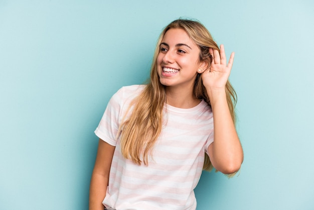 Jonge blanke blonde vrouw geïsoleerd op blauwe achtergrond probeert te luisteren naar een roddel.