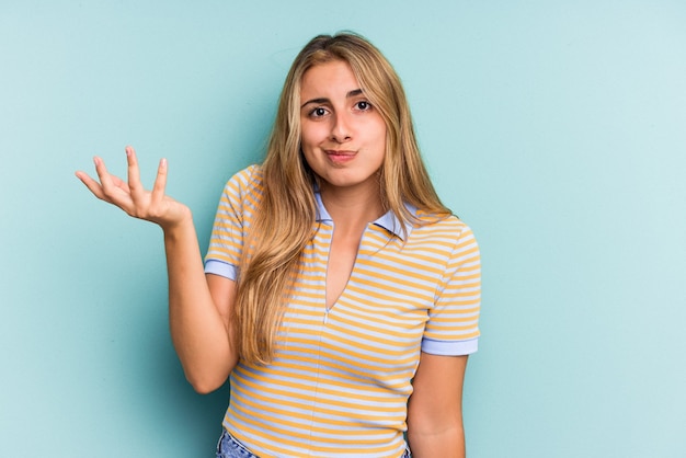 Jonge blanke blonde vrouw geïsoleerd op blauwe achtergrond twijfelen en schouders ophalen in ondervraging gebaar.