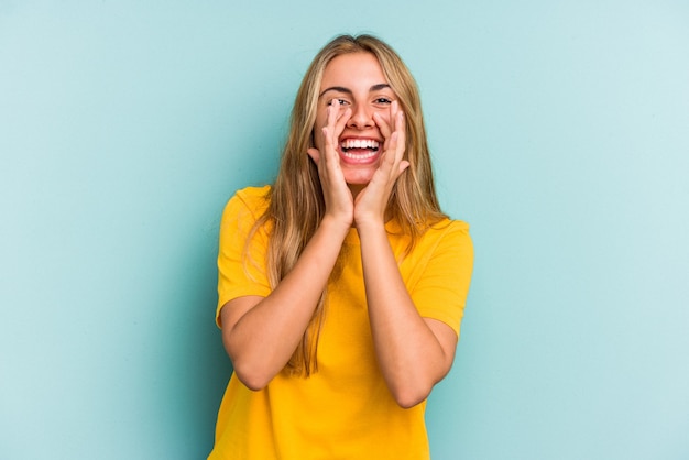 Jonge blanke blonde vrouw geïsoleerd op een blauwe achtergrond die een roddel zegt, wijzend naar de zijkant die iets meldt.