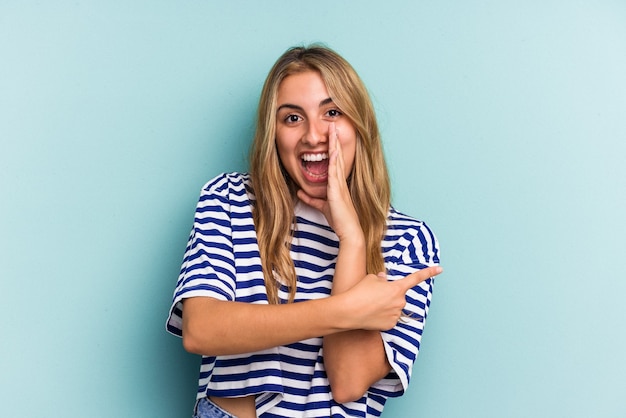 Jonge blanke blonde vrouw geïsoleerd op een blauwe achtergrond die een roddel zegt, wijzend naar de zijkant die iets meldt.