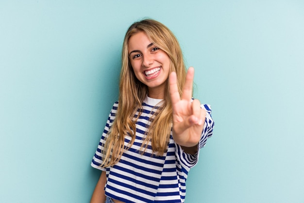 Jonge blanke blonde vrouw geïsoleerd op een blauwe achtergrond die overwinningsteken toont en breed glimlacht.