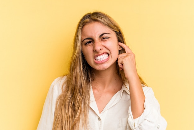 Jonge blanke blonde vrouw geïsoleerd op gele achtergrond die oren bedekt met vingers, gestrest en wanhopig door een luid ambient.