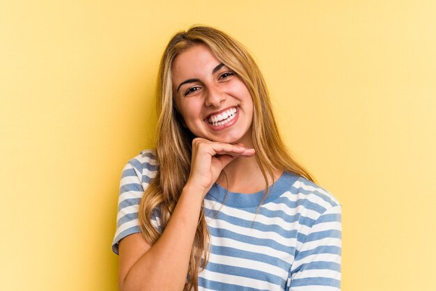 Jonge blanke blonde vrouw geïsoleerd op gele achtergrond glimlachend gelukkig en zelfverzekerd, kin met hand aanraken.