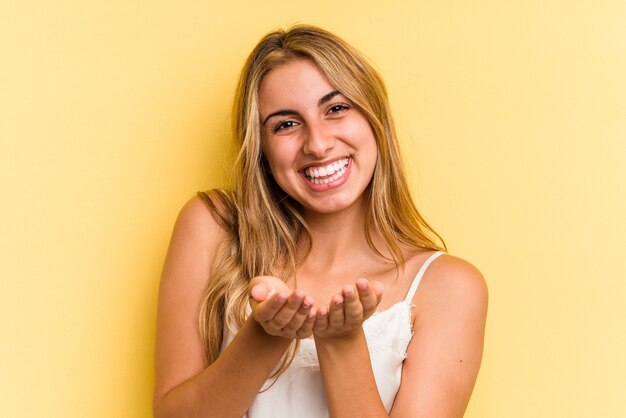 Jonge blanke blonde vrouw geïsoleerd op gele achtergrond met iets met palmen, aanbieden aan camera.