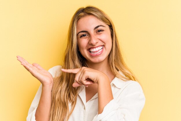 Jonge blanke blonde vrouw geïsoleerd op gele achtergrond opgewonden met een kopie ruimte op palm.