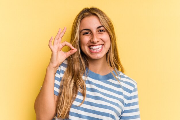 Jonge blanke blonde vrouw geïsoleerd op gele achtergrond vrolijk en zelfverzekerd weergegeven: ok gebaar.