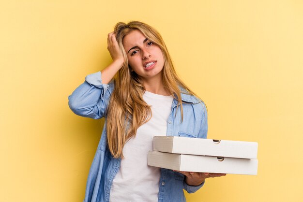 Jonge blanke blonde vrouw met pizza's geïsoleerd op een gele achtergrond die geschokt is, herinnert ze zich een belangrijke vergadering.