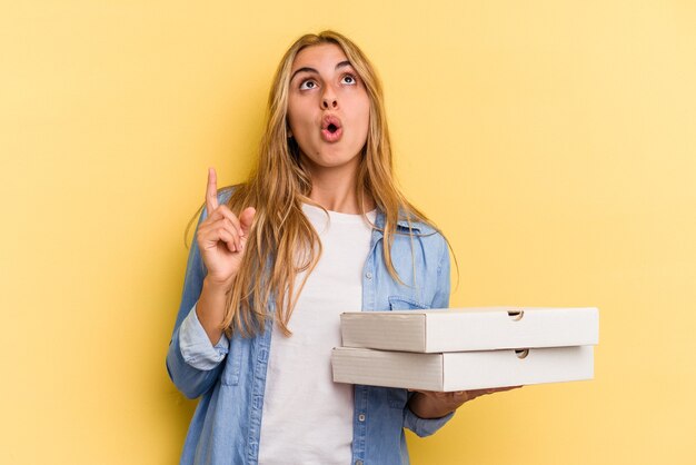 Jonge blanke blonde vrouw met pizza's geïsoleerd op gele achtergrond wijzend ondersteboven met geopende mond.