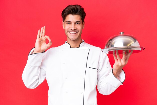 Jonge blanke chef-kok man met dienblad geïsoleerd op rode achtergrond met ok teken met vingers