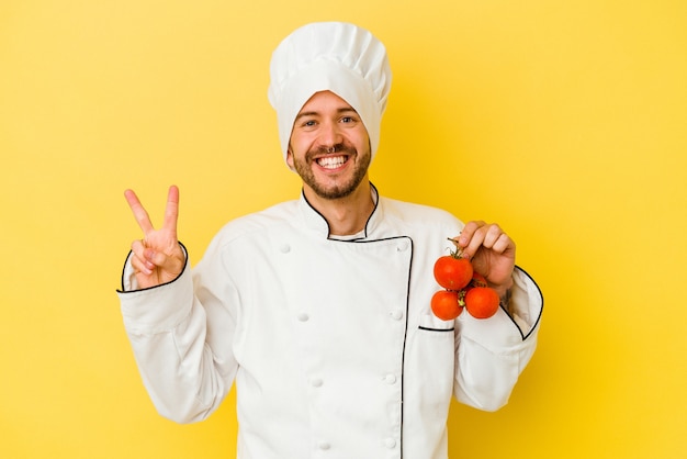 Jonge blanke chef-kok man met tomaten geïsoleerd op gele achtergrond vrolijk en zorgeloos met een vredessymbool met vingers.