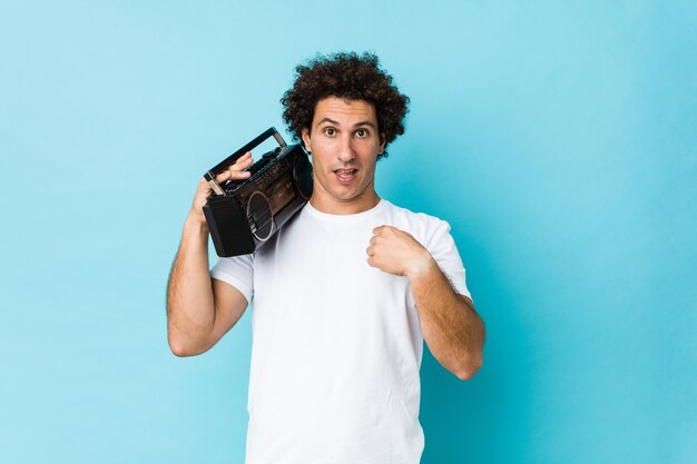 Jonge blanke gekrulde man met een guettoblaster verrast wijzend naar zichzelf, breed glimlachend.