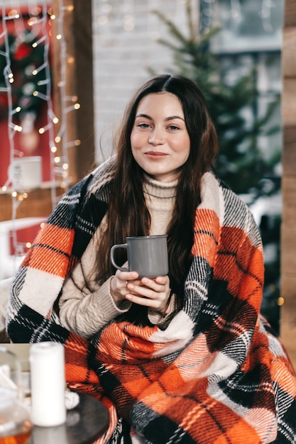 Jonge blanke gelukkige vrouw in een plaid drinkt een warme drank in de achtertuin, genietend van kerstochtend.