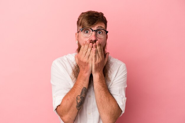 Jonge blanke gember man met lange baard geïsoleerd op roze achtergrond geschokt die mond met handen bedekken.