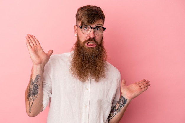 Jonge blanke gember man met lange baard geïsoleerd op roze achtergrond verward en twijfelachtig schouders ophalen om een kopie ruimte te houden.