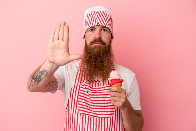 Jonge blanke gember man met lange baard met een ijsje geïsoleerd op roze achtergrond staande met uitgestrekte hand weergegeven: stopbord, voorkomen dat je.