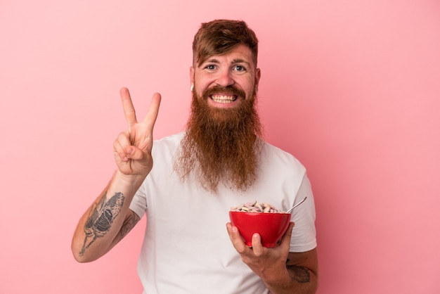 Jonge blanke gember man met lange baard met een kom cornflakes geïsoleerd op roze achtergrond met nummer twee met vingers.
