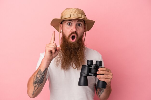 Foto jonge blanke gember man met lange baard met verrekijker geïsoleerd op roze achtergrond met een idee, inspiratie concept.