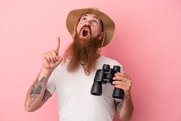 Jonge blanke gember man met lange baard met verrekijker geïsoleerd op roze achtergrond wijzend ondersteboven met geopende mond.