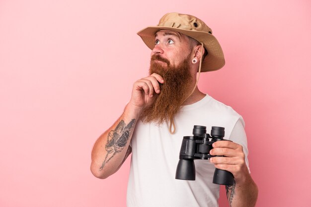 Jonge blanke gember man met lange baard met verrekijker geïsoleerd op roze achtergrond zijwaarts kijkend met twijfelachtige en sceptische uitdrukking.