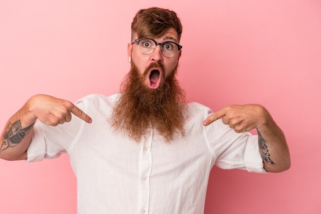 Jonge blanke gemberman met lange baard geïsoleerd op roze achtergrond wijst naar beneden met vingers, positief gevoel.