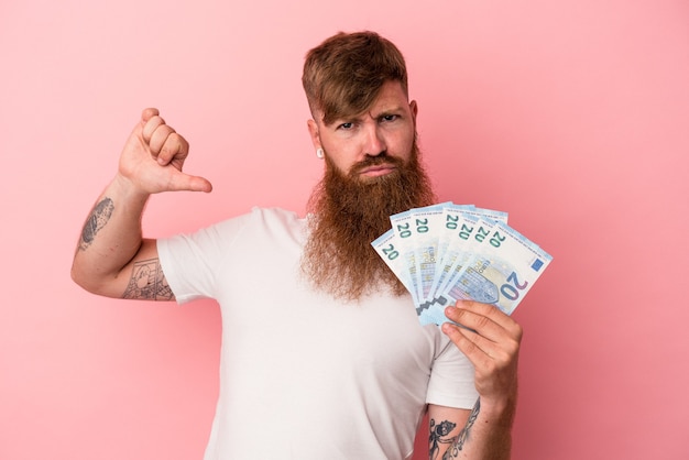 Jonge blanke gemberman met lange baard met bankbiljetten geïsoleerd op roze achtergrond voelt zich trots en zelfverzekerd, voorbeeld om te volgen.