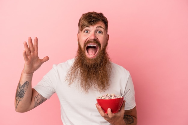 Jonge blanke gemberman met lange baard met een kom cornflakes geïsoleerd op roze achtergrond die een aangename verrassing ontvangt, opgewonden en handen opsteekt.