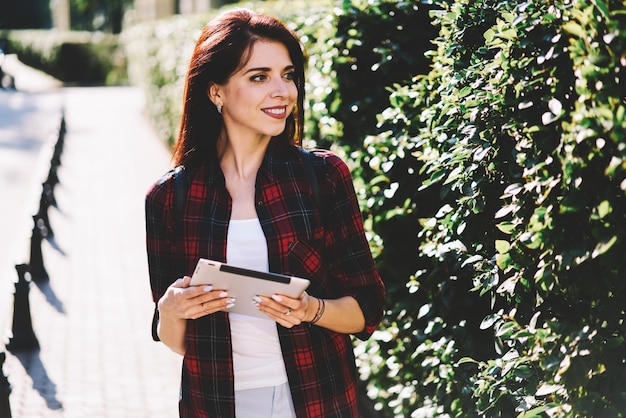 Jonge blanke glimlachende vrouw die opzij kijkt terwijl ze wacht op een vriend op straat met een nieuw touchpad in de hand