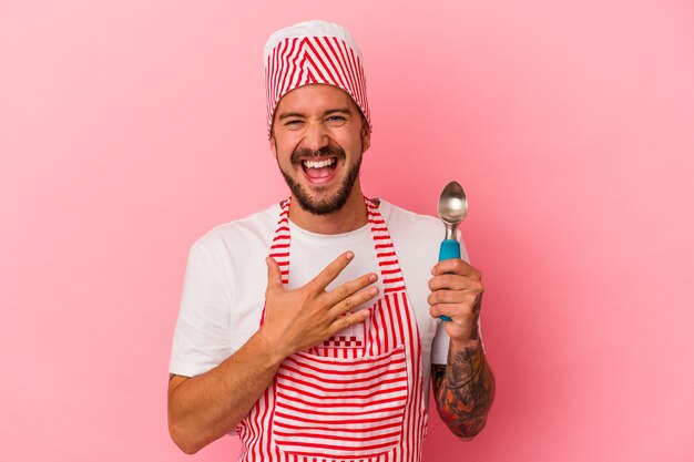 Jonge blanke ijsmaker man met tatoeages met lepel geïsoleerd op roze achtergrond lacht hardop terwijl hij de hand op de borst houdt.