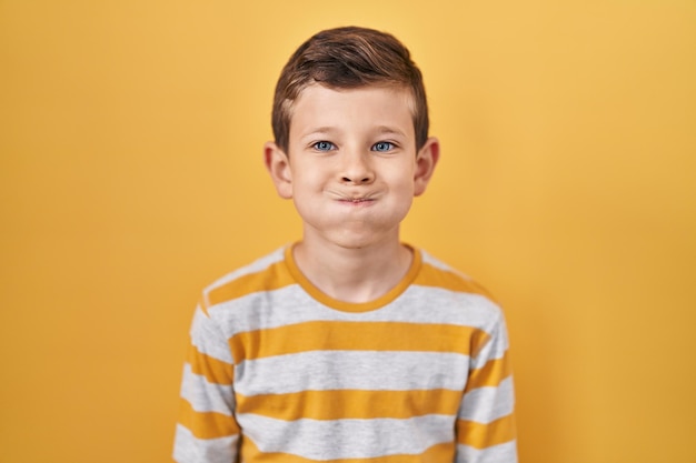 Jonge blanke jongen die op een gele achtergrond staat met opgeblazen wangen met een grappig gezicht, mond opgeblaasd met lucht, gekke uitdrukking.