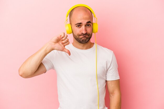 Jonge blanke kale man luisteren naar muziek geïsoleerd op roze achtergrond met een afkeer gebaar, duim omlaag. Onenigheid begrip.