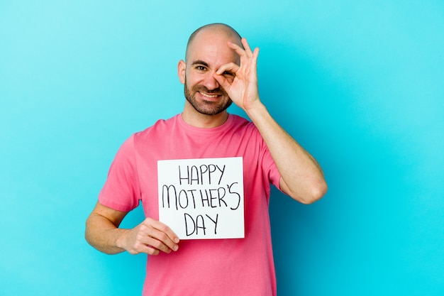 Jonge blanke kale man met een plakkaat met gelukkige moeders dag geïsoleerd op blauw opgewonden houden ok gebaar op oog.
