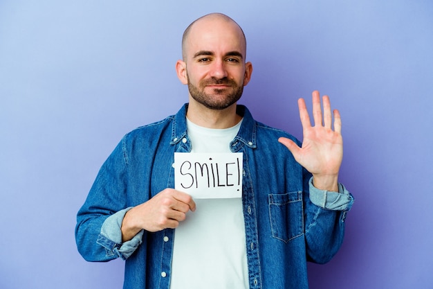 Jonge blanke kale man met een plakkaat Smile geïsoleerd op paarse achtergrond glimlachend vrolijk met nummer vijf met vingers.