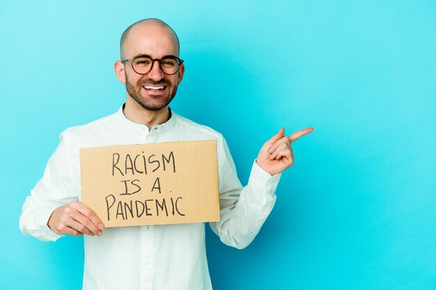 Jonge blanke kale man met een racisme is een pandemie geïsoleerd op een witte muur glimlachend en opzij wijzend, iets laten zien op lege ruimte