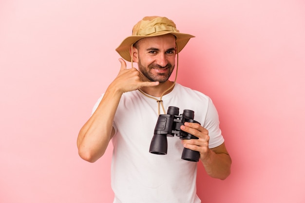 Jonge blanke kale man met verrekijker geïsoleerd op roze achtergrond met een mobiel telefoongesprek gebaar met vingers.