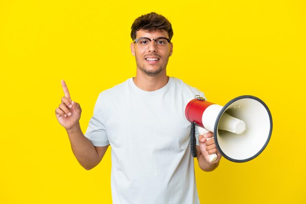 Jonge blanke knappe man geïsoleerd op een gele achtergrond die een megafoon vasthoudt en een geweldig idee naar boven wijst