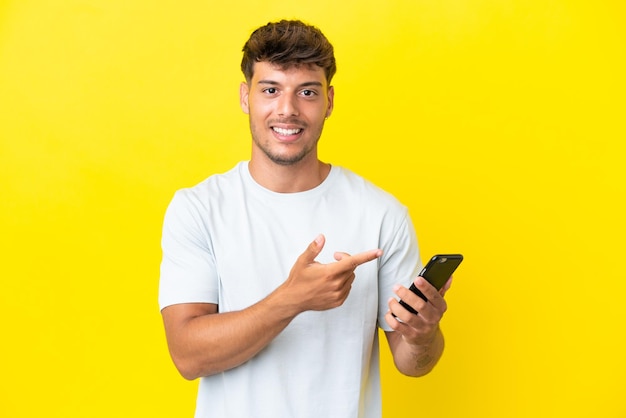 Jonge blanke knappe man geïsoleerd op gele achtergrond met behulp van mobiele telefoon en wijzend