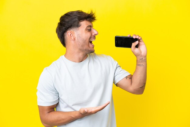 Jonge blanke knappe man geïsoleerd op gele achtergrond met behulp van mobiele telefoon en zingen