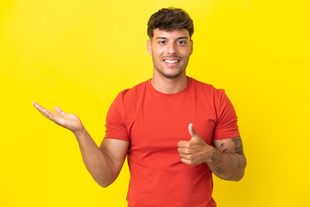 Jonge blanke knappe man geïsoleerd op gele achtergrond met copyspace denkbeeldig op de palm om een advertentie in te voegen en met duimen omhoog