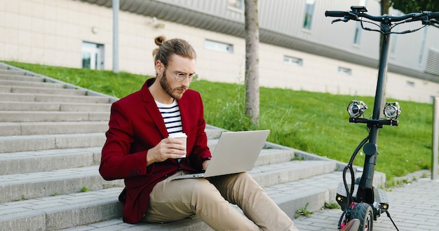 Jonge blanke knappe man in glazen en rode jas zittend op stappen buitenshuis, werken op laptopcomputer, drankje nippen. Stijlvolle man koffie drinken. Freelancer. Elektrische scooter.