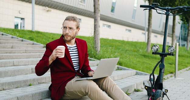 Jonge blanke knappe man in glazen en rode jas zittend op stappen buitenshuis, werken op laptopcomputer, drankje nippen. Stijlvolle man koffie drinken. Freelancer. Elektrische scooter.