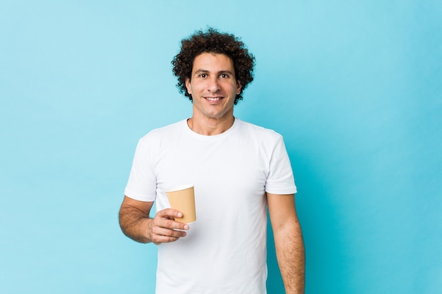 Jonge blanke krullende man met een afhaalmaaltijden koffie blij, glimlachend en vrolijk.