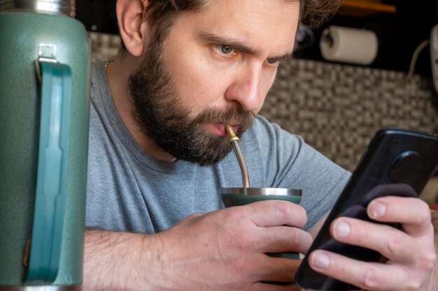 Foto jonge blanke latino man drinkt mate terwijl hij zijn telefoon controleert